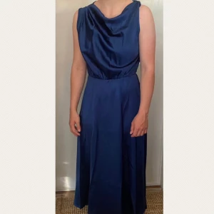 Marinblå långklänning med draperad axel - Oanvänd marinblå långklänning i glansigt material med draperad, asymmetrisk axel och guldfärgad detalj på axelbandet. Klänningen har en markerad midja