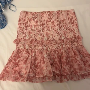 Rosa volangkjol från Gina Tricot - Supersöt kortkjol från Gina Tricot i rosa och vitt med blommigt mönster. Kjolen har smockad midja, volanger och är gjord i ett lätt, flowy material som känns luftigt. Perfekt för dig som gillar romantiska vibes och vill ha något riktigt gulligt i garderoben.