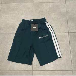 Palm Angels gröna shorts med vita ränder - Palm Angels shorts i mörkgrönt med vita sidränder och logotyp på vänster ben. Modellen har elastisk midja och sportig look, perfekt för chill eller träning. Materialet känns som polyester och passformen är loose.