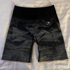 Svarta mönstrade träningsshorts Gymshark - Snygga svarta träningsshorts från Gymshark med grått abstrakt mönster. De har hög midja och är tillverkade i stretchigt syntetmaterial som sitter tight och bekvämt. Perfekta för gymmet eller löpning, med diskret logga framtill.