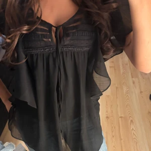 Blus volanger  - Säljer en svart blus från Vila i storlek XS. Blusen har transparenta partier, dekorativa spetsdetaljer och volanger längs sidorna. Den har korta ärmar och knyt framtill vid halsen. Den är knappt använd och som ny