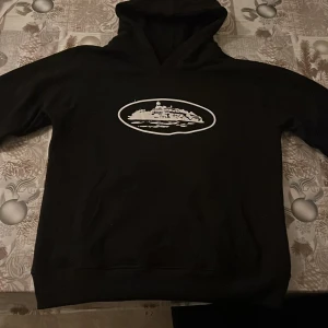 Svart hoodie Cortez med tryck - Svart hoodie från Cortez i storlek S med ett vitt tryck av en byggnad på bröstet. Tröjan har huva och långa ärmar, samt ribbade muddar vid ärmslut och nederkant. Materialet känns mjukt och skönt, perfekt för chill dagar.