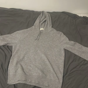 Grå hoodie i kashmir från Sayliss - Mjuk och stilren grå hoodie från Sayliss i lyxig kashmir. Tröjan har huva med snörning, långa ärmar och ribbade muddar vid nederkant och ärmslut. Perfekt för dig som vill ha en chill och snygg look med extra komfort.