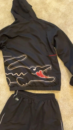 Lacoste tracksuit - Svarta trakcsuit från Lacoste med vita detaljer längs benen och elastisk midja. Materialet är lätt och syntetiskt, perfekt för sport eller chill. Klassisk Lacoste-logga på höften och snygga kontrastlinjer för en modern look. Säljer för har bytt stil. Size L men den sitter som M