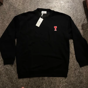 Sweatshirt  - Svart sweatshirt från Ami med klassisk rund hals och lång ärm. På bröstet sitter Ami:s ikoniska broderade hjärta i rött och vitt. Tröjan har ribbade muddar och är tillverkad i mjuk bomull, perfekt för en chill och stilren look.
