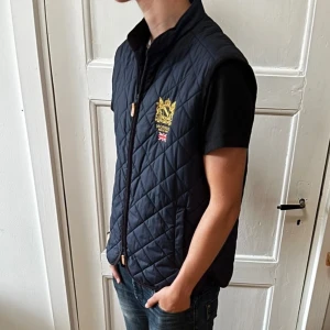 Morris quiltad blå väst med dragkedja - Quiltad marinblå väst från Morris med guldfärgad brodyr och Union Jack-detalj på bröstet. Den är knappt använd och utan skavanker. Storleken är M och personen på bilden 180! Skriv om du har frågor!
