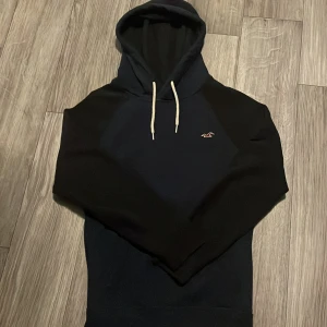 Mörkblå hoodie från Hollister - Säljer en mörkblå hoodie från Hollister med vit snörning och klassisk logga på bröstet. Tröjan har känguruficka framtill och långa ärmar. Perfekt för dig som gillar en avslappnad och stilren look.