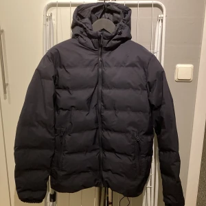 Mörkblå pufferjacka - som ny! - Mörkblå pufferjacka / ”dunjacka” i snyggt matt tyg. Köpt förra året på H&M och knappt använd pga snabbt för liten. Innerficka, två sidofickor med dragkedja och dragsko i jackans nederkant.