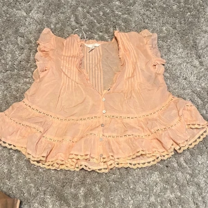 Persikofärgad blus med volangdetaljer - Supersöt persikofärgad blus med volanger och spetsdetaljer längs kanterna. Blusen har v-ringning, knappar framtill och korta volangärmar. Tunt, luftigt tyg som passar perfekt till varmare dagar. Romantisk och feminin vibe. Dock en sönder knapp men den var så när jag köpte den. Inget man tänker på🤗