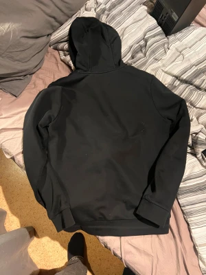 Svart hoodie från BOSS med dragkedja - Svart hoodie från BOSS i storlek M med vit dragkedja och BOSS-logga på bröstet. Tröjan har huva, långa ärmar och två fickor framtill. Passar som S eller Xs