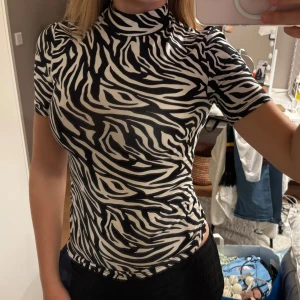 Zebramönstrad topp  - Säljer en topp från amisu med zebramönster. Toppen har korta ärmar och hög krage, tillverkad i stretchigt material som sitter tight mot kroppen. Skriv vid intresse. 