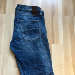 Replay Anbass jeans blå 32 - Snygga Replay Anbass jeans i klassisk blå tvätt med coola slitningar och raka ben. Jeansen har fem fickor, läderpatch bak och tydliga sömmar. Passar dig som gillar en modern och avslappnad stil. Materialet är slitstarkt jeans och midjan är normal.