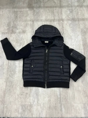 Svart Moncler pufferjacka med huva - Svart pufferjacka från Moncler med stickade ärmar och quiltad kropp. Jackan har huva, två sidofickor med knappar och dragkedja framtill. Moncler-logga på ärmen och ribbade muddar. Perfekt för dig som vill ha en snygg och varm jacka med streetkänsla.       OBS NFC taggen är trasig.