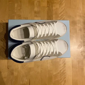 Philippe Model vita sneakers med mockadetaljer - Snygga vita sneakers från Philippe Model med grå mockadetaljer och mörkblå häl. Skorna har klassisk snörning, rund tå och logotyp på sidan. Tillverkade i skinn och mocka för en lyxig känsla. Perfekta för dig som gillar stilrena och trendiga sneakers. pris är diskuterbart 