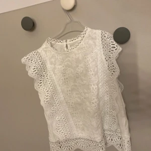 Vit spetsblus med volangdetaljer - Superfin vit blus med broderade spetsdetaljer och volang längs ärmhålen. Blusen är ärmlös och har rund halsringning samt knapp i nacken. Perfekt för dig som gillar romantisk och söt stil. Har ett litet hål i nacken men inget som syns 