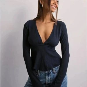 Mörkblå omlottopp från Nelly - Snygg mörkblå omlottopp från Nelly med lång ärm och v-ringad hals. Toppen har ribbad struktur och knytband i midjan som ger en schysst siluett. Perfekt att styla med jeans eller kjol för en cool look.