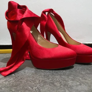 Röda pumps med platå och rosett - Snygga röda pumps med hög klack och platåsula i satinliknande material. långa band som kan knytas runt vristen för en extra statement-look. Perfekta för dig som vill sticka ut med färg och stil.