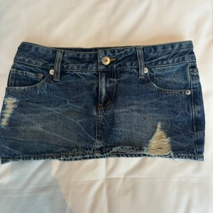 Blå jeanskjol från Cool All, strl 26 - Snygg blå jeanskjol från Cool All i klassisk denim med slitna detaljer och råa kanter. Kjolen har fem fickor, bälteshällor och knäppning fram. Tillverkad i 100% bomull för en autentisk jeanskänsla. Perfekt för dig som gillar en avslappnad och trendig stil.