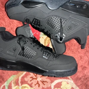 Svarta Nike Jordan 4 sneakers - Säljer ett par svarta Nike Jordan 4 sneakers med ikonisk Jumpman-logga på plösen och hälen. Skorna har meshpaneler, snörning och detaljer i syntetmaterial. Snygg, chunky siluett med platt sula och rund tå. Perfekt för dig som gillar streetwear och basketstil.