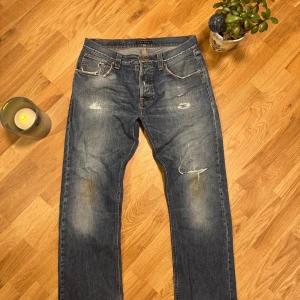 Blå jeans från Nudie Jeans med slitningar - Snygga blå jeans från Nudie Jeans med coola slitningar och lagade detaljer på benen. Klassisk femficksmodell med orangea sömmar och broderade bakfickor. Jeansen har rak passform och är tillverkade i robust denim med vintagekänsla.