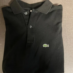Svart pikétröja Lacoste - Klassisk svart pikétröja från Lacoste med den ikoniska gröna krokodilen broderad på bröstet. Tröjan har korta ärmar, krage och två knappar framtill. Materialet är mjuk bomull som andas och passar perfekt till en chill stil. Den är i skick: 10/10, precis som ny! Det är bara att skriva vid minsta fråga, priset är inte hugget i sten!
