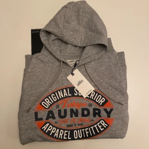  NY Grå hoodie Tokyo Laundry S - Snygg ljusgrå hoodie från Tokyo Laundry i storlek S. Tröjan har huva med snörning och ett stort tryck i orange, svart och vitt på bröstet. Mjuk bomullsmix och klassisk passform, perfekt till jeans. Stilsäker streetwear-vibe med coolt grafiskt tryck. HELT NY