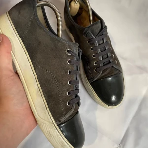  Lanvin - Snygga sneakers från Lanvin i grå mocka med svart glansig tå och vita sulor. Skorna har rund tå, snörning och en stilren design som passar till många outfits. Insidan är i brunt skinn och detaljerna är diskreta men lyxiga.