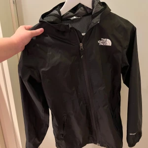 Svart vindjacka från The North Face - Säljer en näst intill helt ny vindjacka från The North Face . Jackan har dragkedja framtill, två sidofickor och logga på bröstet. Materialet är lätt och syntetiskt, perfekt för blåsiga dagar. Snygg och enkel design med lång ärm och justerbar huva. Storlek S junior 