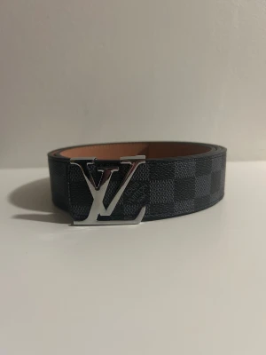 rutig svart bälte - Säljer ett stilrent bälte med klassiskt rutigt mönster i svart och grått. Bältet har en stor logga i silver som spänne och är tillverkat i exklusivt skinn. Perfekt accessoar för att lyfta din outfit.