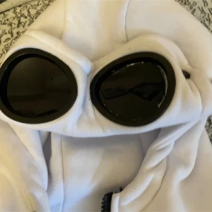 Vit hoodiejacka med goggles från C.P. Company - Säljer en vit hoodiejacka från C.P. Company med ikoniska goggles i huvan och broderad logga på bröstet. Jackan har dragkedja framtill, två stora fickor och är tillverkad i mjuk bomull. Perfekt för dig som gillar streetwear och vill sticka ut med en unik detalj.
