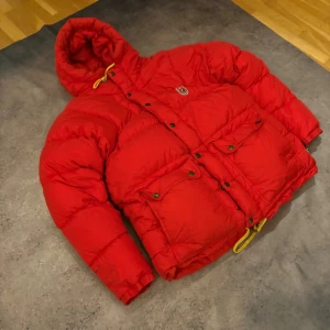 Röd dunjacka från Fjällräven XXS - Säljer en fet röd dunjacka från Fjällräven i storlek XXS. Jackan har huva med gul snörning, två stora fickor med knappar och klassisk Fjällräven-logga på bröstet. Perfekt för kalla dagar och riktigt mysig att bära.