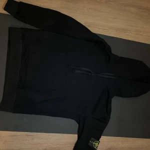 Svart hoodie från Stone Island - Svart hoodie från Stone Island med klassisk logopatch på vänster ärm. Tröjan har huva med snörning, stor magficka och ribbade muddar. Tillverkad i mjuk bomull som är skön att bära. 