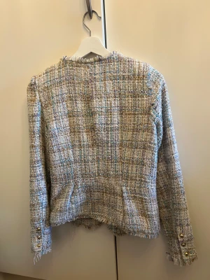 Rutig tweedjacka från Vero Moda - Snygg rutig tweedjacka från Vero Moda i beige, blå och lila toner. Jackan har rund hals, dragkedja framtill och fransiga kanter. Guldknappar på ärmsluten ger extra stilpoäng. Perfekt för dig som gillar en chic och trendig look.
