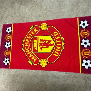 Manchester United halsduk röd/gul - Stor och färgstark halsduk med Manchester Uniteds ikoniska klubbmärke i gult och rött. Kanterna har fotbollsmönster och mindre klubbmärken. Perfekt för dig som vill visa ditt stöd på matchen eller hemma. Materialet är mjukt och passar bra runt halsen.