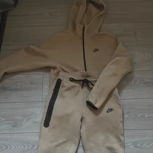Beige mjukisbyxor från Nike - Äkta sett från Nike med svart logga på benet och på bröstet. De är inte så använda, skicket är inprincip nytt. Byxorna är i strl S och koftan i strl XS (man storlek) hör av er vid frågor. 1200kr för både byxorna och koftan 🤩