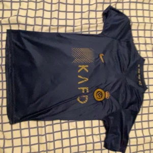 Blå Ronaldo fotbollströja Nike - Snygg marinblå fotbollströja från Nike med guldiga detaljer och tryck. Framsidan har klubbmärke och sponsorlogga, medan baksidan har Ronaldo och nummer 7 i guld. Kortärmad och tillverkad i lätt, ventilerande material som passar perfekt för träning eller match.