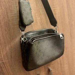 svart axelväska med monogram mönster  - Snygg svart axelväska med klassiskt monogrammönster. Väskan har flera fack, silverfärgade dragkedjor och bred justerbar axelrem med logga. Tillverkad i skinn och levereras med matchande liten pouch och extra ficka.