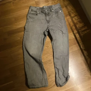 Grå jeans från Jack & Jones  - Säljer ett par grå jeans från Jack & Jones, modellen Relaxed Chris. De har en avslappnad passform med raka ben och klassisk femficksdesign. Jeansen är tillverkade i bomull och har silverfärgade detaljer vid knappar och nitar. Perfekt för en chill och stilren look.