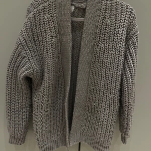 Beige stickad kofta med grov struktur - Säljer en beige stickad kofta med grov struktur och breda ribbade muddar. Koftan har lång ärm och öppen framtill, perfekt för lager på lager. Den har en loose passform och känns riktigt mysig att dra på sig när det är kyligt.