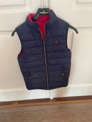 Vändbar dunväst från Polo Ralph Lauren - Tjena! Vändbar dunväst från Polo Ralph Lauren med ena sidan marinblå och andra sidan röd. Västen har dragkedja, ståkrage och två fickor med knappstängning på båda sidor. Ikoniska Polo-loggan broderad på bröstet. Perfekt för lager-på-lager och snygg streetstil. Skick: 8/10. Storlek: xs. Bara att fråga om det är något! Priset går att diskutera!😊