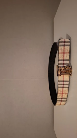 Stilig bälte med guld spänne 2 sidig - Säljer ett snyggt 2 sidigt med klassiskt rutigt mönster i beige, svart och rött. Bältet har ett exklusivt guldigt spänne med  ikoniska TB-logga och är tillverkat i skinn. Perfekt accessoar för att lyfta din outfit med lyxig känsla.