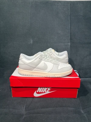Nike Dunk Low i ljusgrått skinn - Helt nya och oanvända Nike Dunk Low sneakers i ljusgrått och vitt skinn med klassisk rund tå och platt sula. Snygg snörning och perforerade detaljer på tån för extra stil. Sulan har en lätt beige ton som ger en fräsch kontrast. Perfekt för dig som gillar cleana och stilrena sneakers.