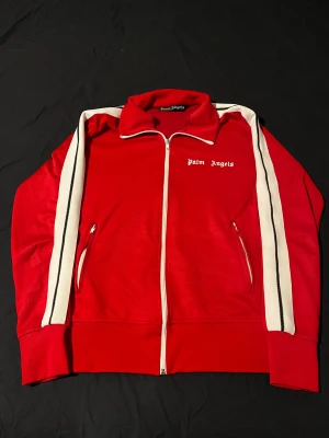 Röd track jacket från Palm Angels - Palm Angels röd track jacket med vita och svarta ränder längs ärmarna, klassisk ståkrage och hel dragkedja framtill. Jackan har två fickor med dragkedja och vit Palm Angels-logga på bröstet. Materialet är mjuk polyester med sportig vibe. Hör av er vid frågor!