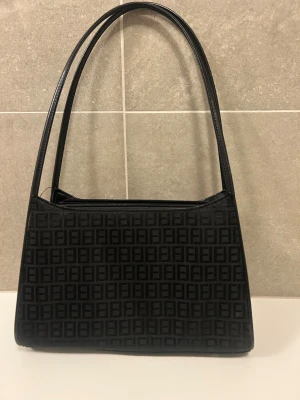 Svart Fendi axelväska med FF-mönster - Snygg svart axelväska med klassiskt FF-mönster över hela väskan. Väskan har en stilren rektangulär form, långa handtag och dragkedja upptill samt en extra ficka med dragkedja på baksidan. Mått: 29 x 20 x 9 cm