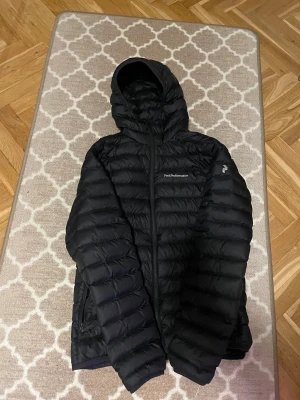 Svart dunjacka Peak Performance - Svart dunjacka från Peak Performance med huva och snyggt quiltad design. Jackan har dragkedja framtill och logga på bröstet samt ärmen. Perfekt för kalla dagar och har en clean, sportig look som funkar till streetwear.