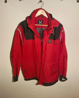 Röd vindjacka från Haglöfs med huva - Snygg röd vindjacka från Haglöfs med svarta detaljer och reflexer på axlarna. Jackan har huva, meshfoder, dragkedja och tryckknappar framtill samt justerbara muddar. Perfekt för dig som vill ha en funktionell och stilren jacka till vardags.