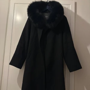 Svart kappa med pälskrage från Hollies - Jättefin kappa i 100% ull, innerfoder 100% polyester. Fin svart päls på huvan. Medföljer skärp i samma material för att knyta runt midjan. En knapp har lossnat för att knäppa kappan på insidan (se bild 5). men extra knapp finns i etiketten och enkelt att sy på vid önskemål. Knapp finns vid halsen! En tuggumifläck finns på insidan av kappan (torr).