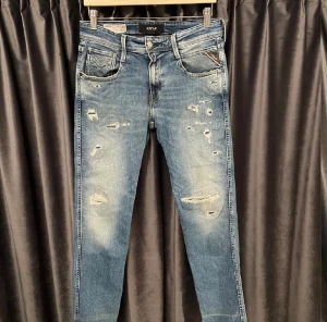 Replay Anbass blå slitna jeans - Snygga Replay Anbass jeans i blå tvätt med slitningar och distressed detaljer på benen. Klassisk femficksmodell med logotyp på bakfickan och läderpatch i midjan. Jeansen har en rak passform och är gjorda i mjukt denimtyg för en cool look.
