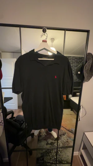 Svart pikétröja från Polo Ralph Lauren - Klassisk svart pikétröja från Polo Ralph Lauren med röd broderad logga på bröstet. Tröjan har korta ärmar, krage och knappar vid halsen. Perfekt för en clean och stilren look, passar lika bra till jeans som till shorts.