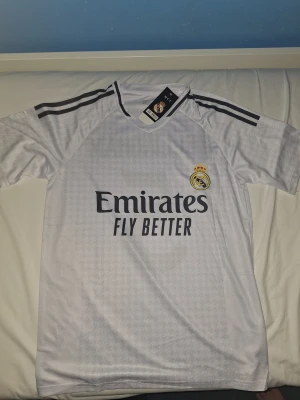 Real Madrid Bellingham #5 fotbollströja - Säljer en vit Real Madrid fotbollströja från Adidas med Bellingham och nummer 5 på ryggen. Tröjan har svarta ränder på axlarna, klubbmärke på bröstet och Emirates Fly Better-tryck. Tillverkad i lätt och ventilerande polyester, perfekt för match eller träning.